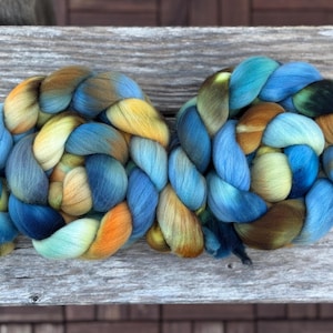 Peut inclure: Deux écheveaux de fil teints à la main, tressés. Le fil présente un mélange de bleus, de jaunes et d'oranges, créant un effet de dégradé. Le fil est présenté sur une surface en bois vieilli.