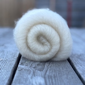 May include: A tightly rolled ball of natural, off-white wool, showcasing a spiral pattern. The wool appears soft and fluffy, with a slightly textured surface. The image is taken on a weathered, gray wooden surface.
