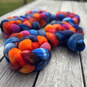 May include: Braided roving yarn in vibrant colors of blue, orange, pink, and purple. The yarn is arranged in a circular pattern, showcasing the blend of hues. The fiber is soft and fluffy, perfect for knitting or other fiber arts projects.
