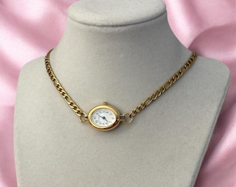 May include: Gold-toned necklace featuring an oval watch face with a white dial and black numerals. The watch is connected to a chain with a textured, linked design. The necklace is displayed on a mannequin against a pink background.