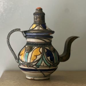 May include: A traditional Moroccan teapot with a silver lid and handle. The teapot is decorated with blue, green, yellow, and white geometric patterns.