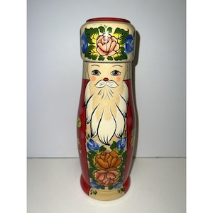 May include: A hand-painted wooden Santa Claus figurine. The figurine has a red coat, a white beard, and a hat decorated with flowers. The base is a light beige colour. The figurine is approximately 20 cm tall.