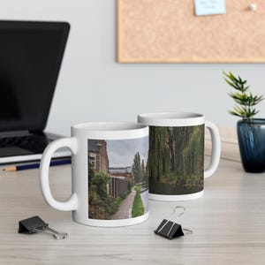 Op de afbeelding: Twee witte keramische mokken met een fotoprint van een pad langs een kanaal, omzoomd door bakstenen gebouwen en weelderige groene treurwilgen. De mokken hebben een standaard cilindrische vorm met een gebogen handvat en een landschapsfoto.