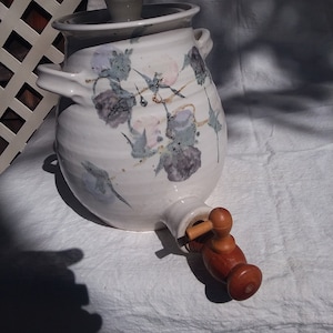 May include: White ceramic pitcher with a floral design and a wooden spigot. The pitcher has a lid with a small knob on top.