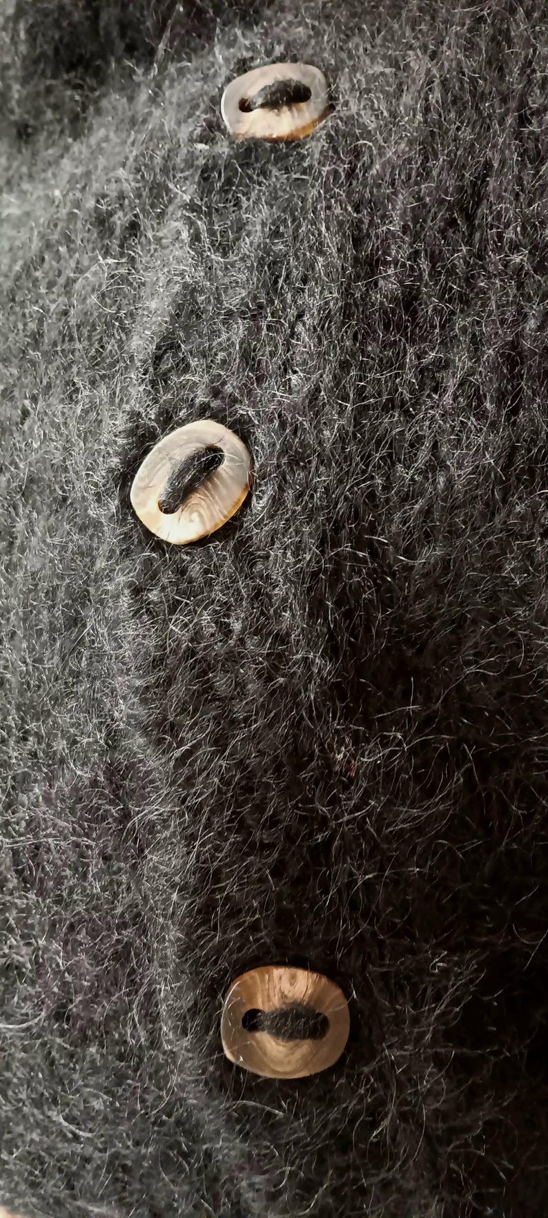 Op de afbeelding: Close-up van een donkergrijze, getextureerde stof met drie ovale knopen. De knopen hebben een lichtbruine, parelmoerachtige uitstraling en zijn gelijkmatig over de stof verdeeld.