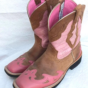May include: A pair of brown and pink cowboy boots. The boots have a square toe and a pink vamp with decorative cutouts. The uppers are brown with pink accents and a pink interior. The boots have a black sole and a brown pull tab.