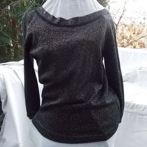 May include: A black long-sleeved sweater with a sparkly, textured finish. The sweater has a wide, boat-neck style neckline with a black trim. The garment is displayed on a white mannequin, showcasing its shape and design. The sweater's fabric appears to have a subtle shimmer.