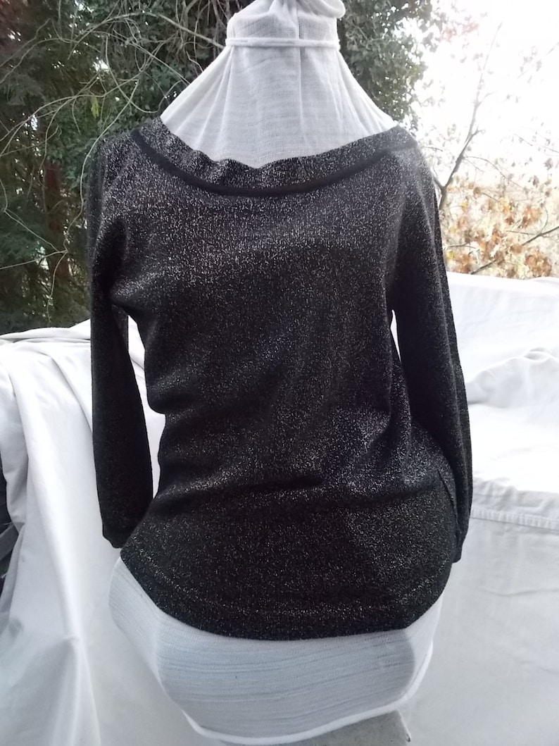 May include: A black, long-sleeved sweater with a sparkly texture. The boat-neck neckline is trimmed with black. The garment is displayed on a white mannequin, highlighting its shape and design. The fabric of the sweater has a subtle shimmer, giving it a textured appearance.