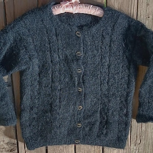 Op de afbeelding: Een donkerblauwe cardigan met knopen en een textuurgebreid patroon. De trui met lange mouwen heeft een ronde hals en wordt op een roze hanger gepresenteerd. De cardigan heeft zes knopen aan de voorkant, op een houten achtergrond.