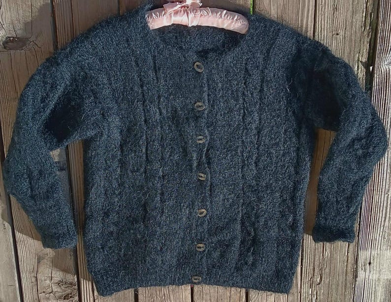Op de afbeelding: Een donkerblauwe cardigan met knopen en een textuurgebreid patroon. De trui met lange mouwen heeft een ronde hals en wordt op een roze hanger gepresenteerd. De cardigan heeft zes knopen aan de voorkant, op een houten achtergrond.