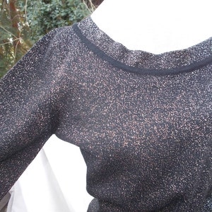 May include: A black long-sleeved sweater with a round neckline. The sweater has a subtle metallic shimmer throughout, with flecks of gold or bronze. The neckline and cuffs are trimmed with a solid black band. The fabric appears to be a knit.
