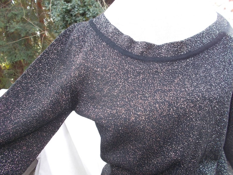 May include: A black long-sleeved sweater with a round neckline. The sweater has a subtle metallic shimmer throughout, with flecks of gold or bronze. The neckline and cuffs are trimmed with a solid black band. The fabric appears to be a knit.