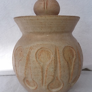 May include: A beige ceramic jar with a lid. The jar has a textured surface with a repeating pattern of rounded shapes. The lid is a smooth, rounded sphere.