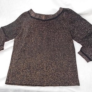 May include: A long-sleeved top with a textured, speckled pattern in shades of brown and gold. The neckline and sleeve cuffs are trimmed with a thin black band. The top has a scoop neckline and a slightly flared shape.
