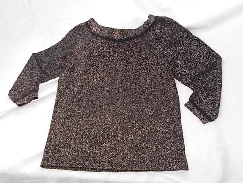 May include: A long-sleeved top with a textured, speckled pattern in shades of brown and gold. The neckline and sleeve cuffs are trimmed with a thin black band. The top has a scoop neckline and a slightly flared shape.