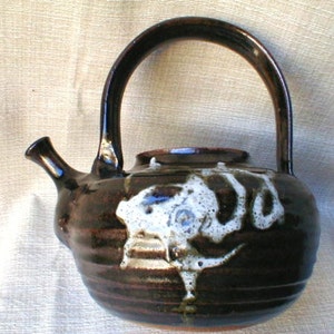 May include: A handcrafted ceramic teapot with a dark brown glaze and a white abstract design. The teapot has a curved handle and a spout. The design is reminiscent of a cloud. The teapot is a unique piece of kitchenware.