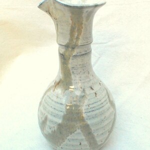 Può includere: Un'anfora in ceramica bianca con un'ampia apertura e un corpo arrotondato. L'anfora ha una smaltatura grigio chiaro con striature di una smaltatura grigio più scuro.