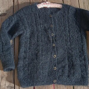 Op de afbeelding: Een donkerblauwe cardigan met knopen en een textuur breipatroon. De trui heeft lange mouwen en een ronde halslijn. De knopen zijn lichtbruin. De trui ligt op een houten ondergrond.