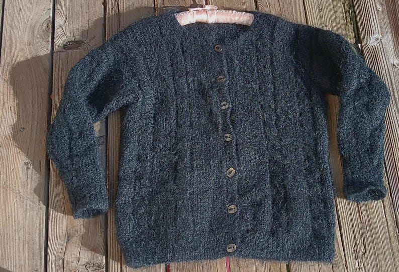 Op de afbeelding: Een donkerblauwe cardigan met knopen en een textuur breipatroon. De trui heeft lange mouwen en een ronde halslijn. De knopen zijn lichtbruin. De trui ligt op een houten ondergrond.