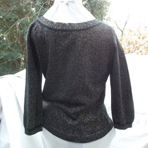 May include: Black sweater with gold metallic thread, featuring a scoop neckline and three-quarter sleeves. The garment has a fitted waist and is displayed on a white mannequin. The background is blurred.