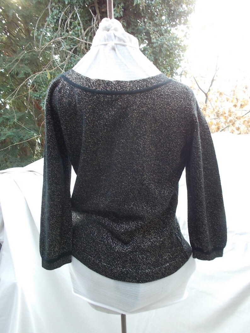 May include: Black sweater with gold metallic thread, featuring a scoop neckline and three-quarter sleeves. The garment has a fitted waist and is displayed on a white mannequin. The background is blurred.