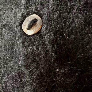 Op de afbeelding: Close-up van een donkergrijze, fuzzy stof met drie ovaalvormige knopen. De knopen hebben een lichtbruine, parelmoerachtige uitstraling met een donker midden. De stof lijkt een trui of een soortgelijk kledingstuk te zijn.