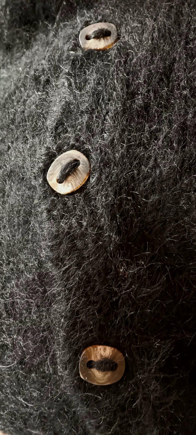 Op de afbeelding: Close-up van een donkergrijze, fuzzy stof met drie ovaalvormige knopen. De knopen hebben een lichtbruine, parelmoerachtige uitstraling met een donker midden. De stof lijkt een trui of een soortgelijk kledingstuk te zijn.