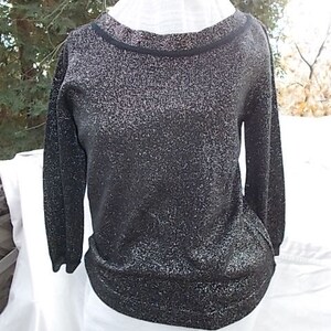 May include: A black, long-sleeved sweater with a sparkly, glittery texture. The sweater has a round neckline with a black trim. The bottom hem and cuffs are also black. The sweater is displayed on a white form.