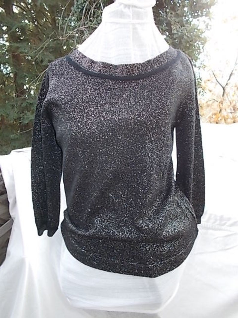 May include: A black, long-sleeved sweater with a sparkly, glittery texture. The sweater has a round neckline with a black trim. The bottom hem and cuffs are also black. The sweater is displayed on a white form.