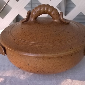 May include: A brown ceramic casserole dish with a lid and two handles. The dish has a speckled glaze and a slightly rounded shape.