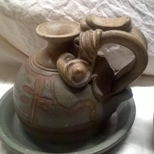 May include: A handcrafted ceramic pitcher and saucer set. The pitcher is a muted green and brown with a decorative handle and spout. The saucer is a matching muted green. The set is on a white background.