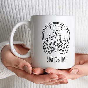 May include: White ceramic mug with a black line art design featuring headphones, flowers, and a cloud with rain. The words "STAY POSITIVE" are printed in black below the design. The mug is held by a person.