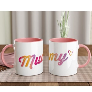May include: Two white ceramic mugs with pink interiors and handles. The word "Mummy" is written on each mug in a colorful, gradient font. One mug has a small heart symbol next to the word.