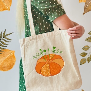 Könnte beinhalten: Beige Tragetasche mit einem orangefarbenen Kürbismotiv und grünen Akzenten. Der Kürbis hat einen Knopf in der Mitte und ist mit verschiedenen Mustern verziert. Die Tasche hat cremefarbene Griffe und wird von einer Person in einem grünen Polka-Dot-Kleid gehalten.
