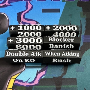 May include: Black rectangular game tokens with white text, including numbers and phrases like "Blocker" and "Double Atk." The tokens are stacked on a colourful playmat with a cartoon character design.