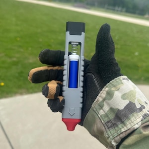 May include: A grey and red 3D-printed device with a blue cylindrical component. The device has a spring mechanism and a black top. The image is taken outdoors, with a green grassy background.