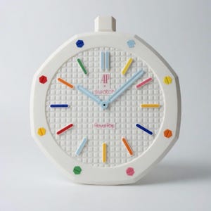 Könnte beinhalten: Weiße achteckige Uhr mit strukturiertem Zifferblatt und farbigen Stundenmarkierungen. Die Uhr hat hellblaue Zeiger und den Text "AP x Swatch Royal Pop". Die Uhr hat eine quadratische Oberseite.