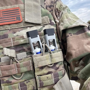 Peut inclure: Gros plan d'un gilet tactique de camouflage avec deux dispositifs gris et noirs fix&eacute;s dans des poches avant. Un patch du drapeau am&eacute;ricain est visible. Le gilet est con&ccedil;u pour un usage militaire ou tactique.
