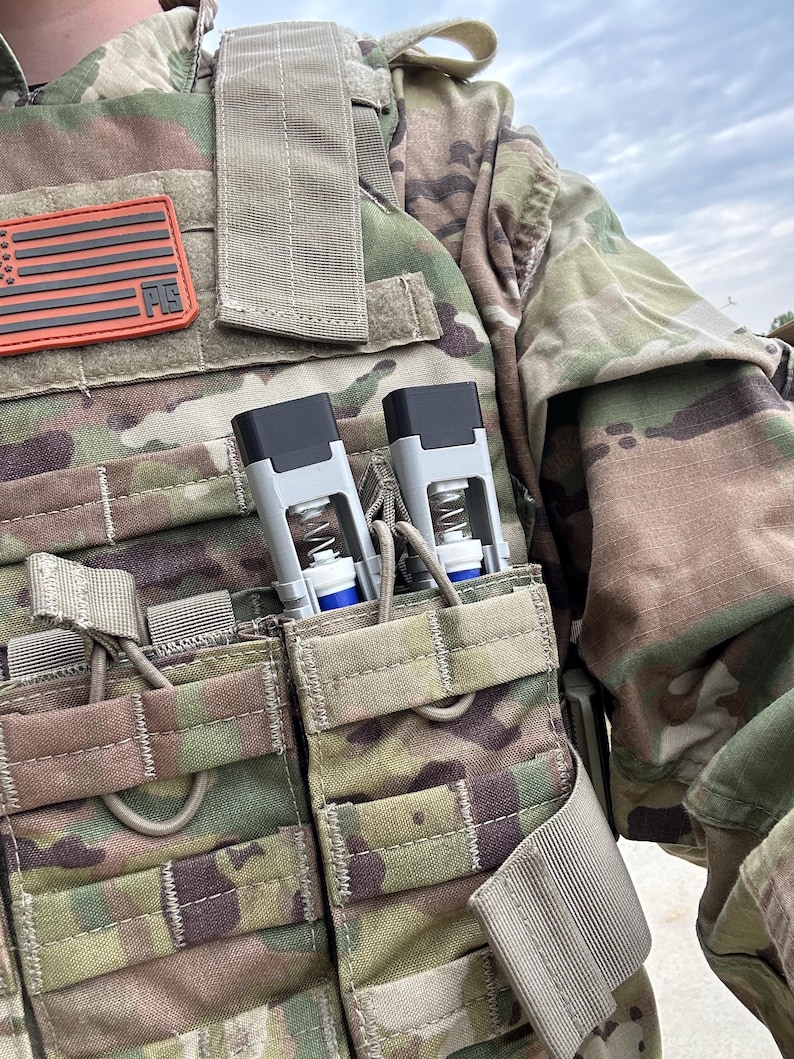 Peut inclure: Gros plan d'un gilet tactique de camouflage avec deux dispositifs gris et noirs fix&eacute;s dans des poches avant. Un patch du drapeau am&eacute;ricain est visible. Le gilet est con&ccedil;u pour un usage militaire ou tactique.