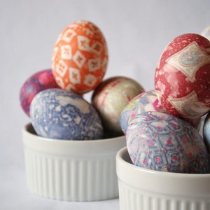 May include: A collection of decorated Easter eggs in white ceramic ramekins. The eggs feature various patterns and colors, including orange with white diamond shapes, blue with floral designs, and red with geometric patterns. A festive and colorful display.