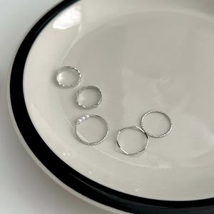May include: Five silver rings of different designs, including beaded, textured, and smooth bands. One ring has three small pearls. The rings are arranged on a white plate with a black rim, creating a minimalist aesthetic.
