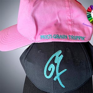 Könnte beinhalten: Zwei Baseballkappen, eine rosa und eine schwarze. Die rosa Kappe hat den Text "MULTI-GRAIN TRIPP'N" auf der Vorderseite gestickt. Die schwarze Kappe hat ein türkisblaues Grafik der Buchstaben "G" auf der Vorderseite. Beide Kappen haben einen gebogenen Schirm.