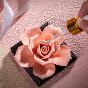 May include: A ceramic rose flower diffuser with a black base. A glass dropper bottle with a gold cap is being used to add essential oil to the diffuser.
