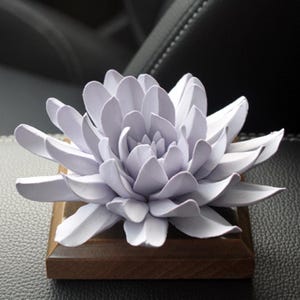 May include: A pale purple flower sculpture with layered petals, set on a small, square, wooden base. The flower is the main focus, with a detailed, realistic design. The wooden base provides a contrasting, natural element.