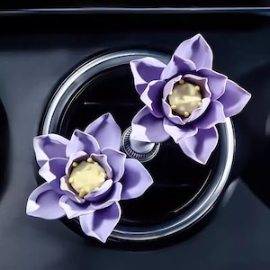 May include: Two purple flower-shaped car air fresheners with yellow centres. The air fresheners are attached to a black car vent.