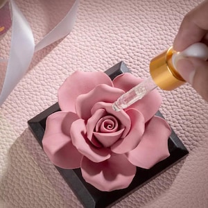 Könnte beinhalten: Eine rosa Keramikrose mit schwarzem Sockel, auf die ätherisches Öl aus einer Glaspipettenflasche mit goldener Kappe getropft wird.