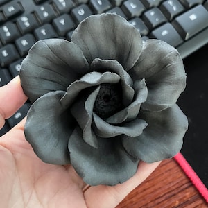 May include: A dark gray, handcrafted flower with multiple petals. The flower is held in a hand, with a keyboard in the background. The flower's center is a darker shade of gray, creating a contrast with the petals.