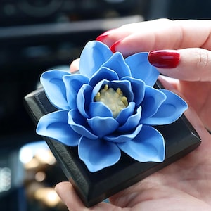 Può includere: Un fiore di ceramica blu con un centro giallo, seduto su una base quadrata nera. Il fiore è un design di loto.