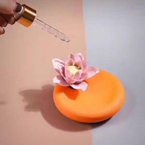 Może przedstawiać: Okrągły, pomarańczowy ceramiczny talerz z różowym i żółtym kwiatem na wierzchu. Przezroczysty szklany zakraplacz ze złotą nasadką jest trzymany nad talerzem, z kroplą cieczy spadającą. Tło jest podzielone na różowe i szare.