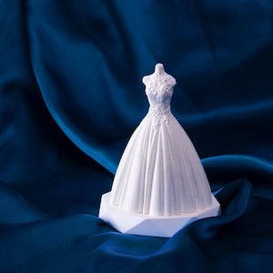 May include: A white porcelain figurine of a wedding dress on a white pedestal base. The figurine is displayed on a dark blue fabric background.